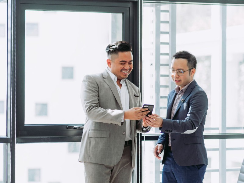 Two men in business attire smiling while looking at a mobile device