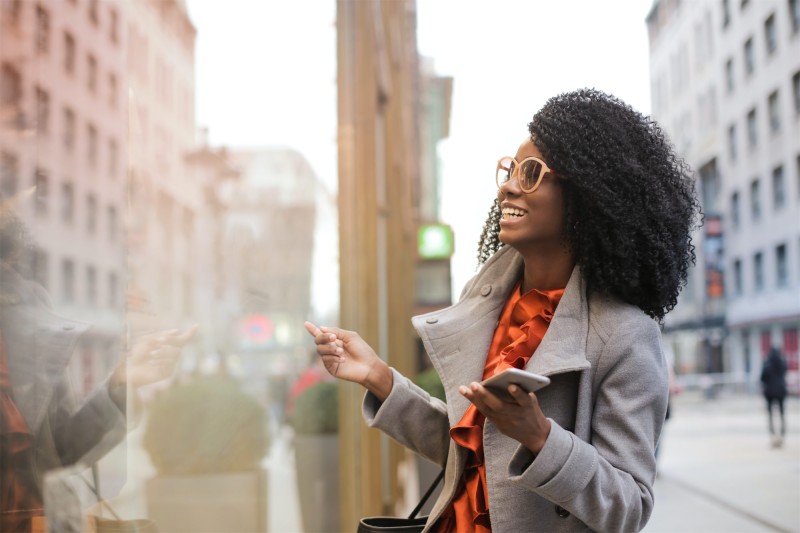 A woman holding a mobile device speaking with someone through a glass window